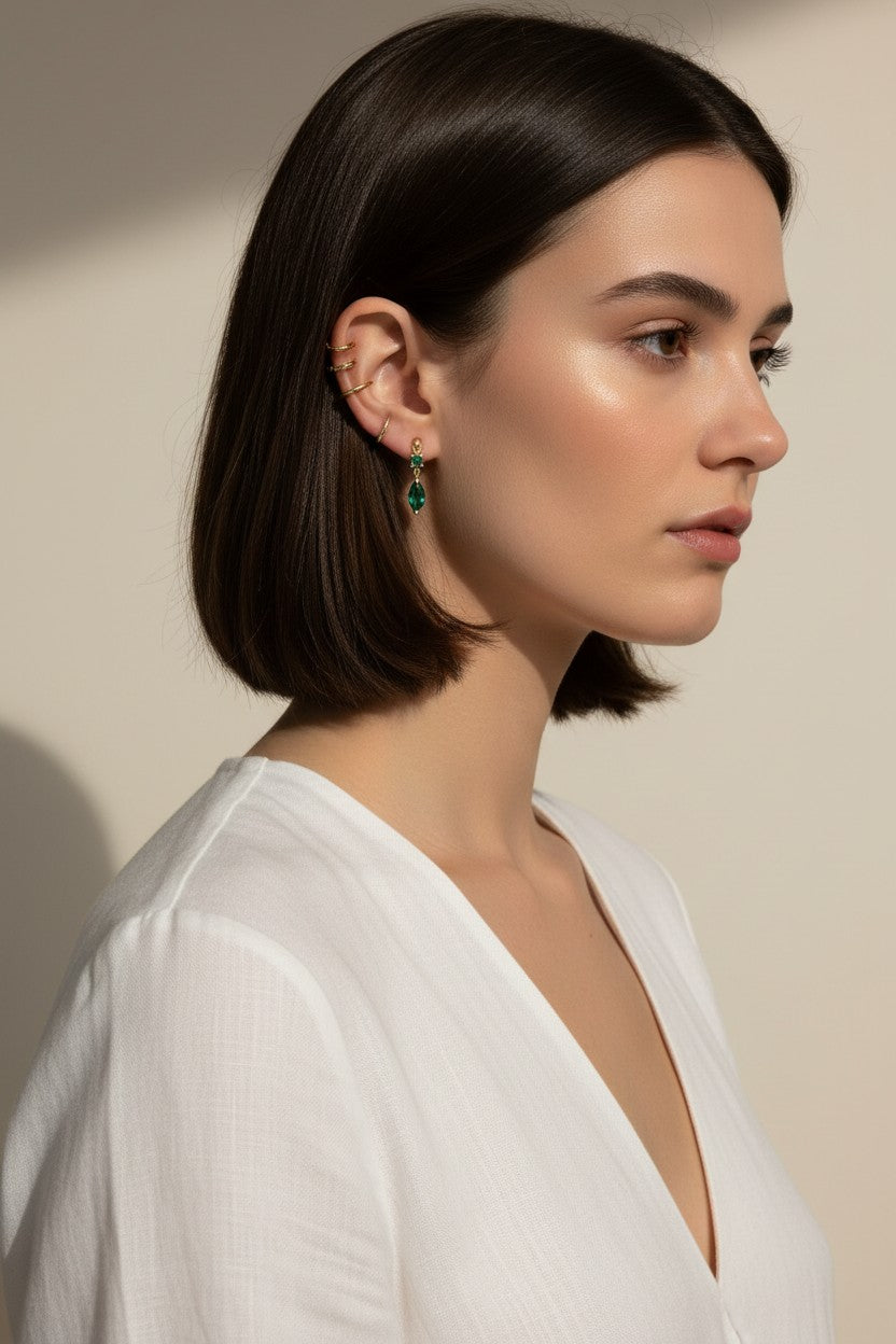 This close-up profile shot shows the emerald green marquise drop earring worn on the lobe, its faceted stone catching soft light for a rich, jewel-toned sparkle. Styled with a sleek center-part bob and layered with multiple thin gold ear cuffs along the upper ear, the earring is shown at true scale, giving shoppers a clear sense of its drop length and how it moves against the jawline. The warm, dramatic side lighting and minimalist white top create a polished, editorial mood, highlighting how the emerald dr