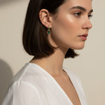 This close-up profile shot shows the emerald green marquise drop earring worn on the lobe, its faceted stone catching soft light for a rich, jewel-toned sparkle. Styled with a sleek center-part bob and layered with multiple thin gold ear cuffs along the upper ear, the earring is shown at true scale, giving shoppers a clear sense of its drop length and how it moves against the jawline. The warm, dramatic side lighting and minimalist white top create a polished, editorial mood, highlighting how the emerald dr