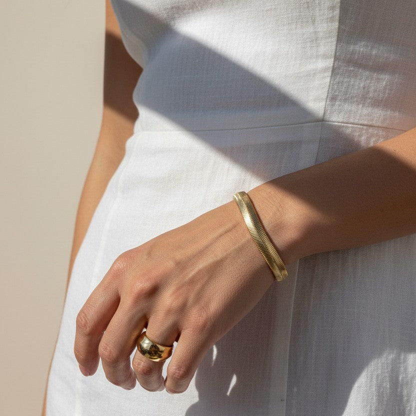 This sunlit close-up shows a gold herringbone chain bracelet wrapped around the wrist, its smooth, flat links catching warm natural light for a rich, reflective shine. Styled with a crisp white linen dress and a chunky gold dome ring on the finger, the bracelet is shown at true scale, giving shoppers a clear sense of its fit and how it drapes against the skin. Bold, graphic shadows and golden-hour lighting create a striking editorial mood, while the relaxed hand pose highlights how the sleek herringbone tex