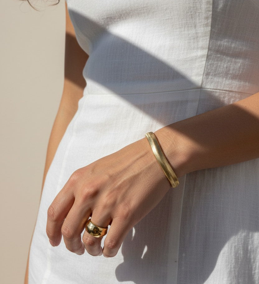 This sunlit close-up shows a gold herringbone chain bracelet wrapped around the wrist, its smooth, flat links catching warm natural light for a rich, reflective shine. Styled with a crisp white linen dress and a chunky gold dome ring on the finger, the bracelet is shown at true scale, giving shoppers a clear sense of its fit and how it drapes against the skin. Bold, graphic shadows and golden-hour lighting create a striking editorial mood, while the relaxed hand pose highlights how the sleek herringbone tex