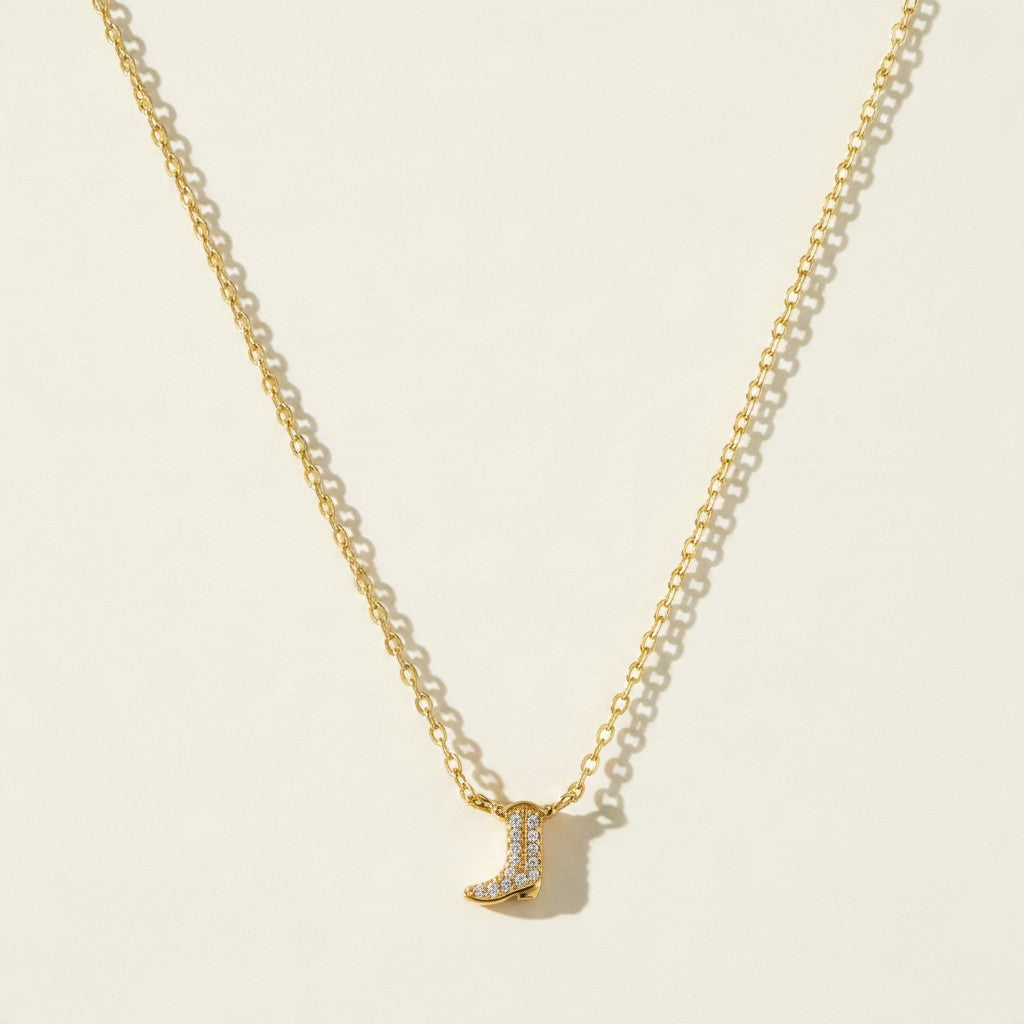 This dainty necklace has a tiny cowboy boot charm in polished gold-tone metal, covered in rows of small clear stones set in a pavé pattern. It hangs from a fine cable chain. The first frame shows the clasp end: a spring ring clasp with an adjustable extender of open curb-style links ending in a tiny stone-set drop. The second is an angled close-up of the pavé boot charm sitting on the chain in soft light. The third lays the whole necklace in a V shape on a warm cream surface, with the boot centered at the b