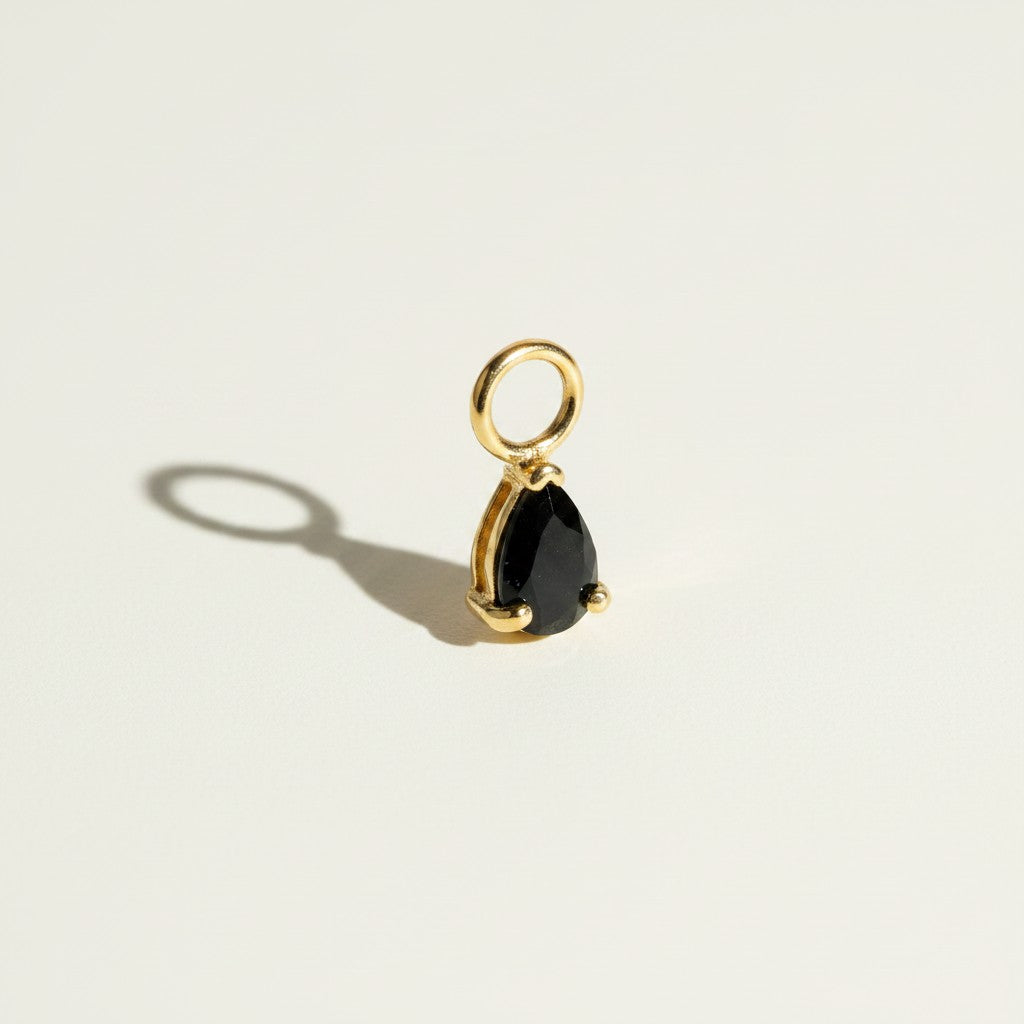 These interchangeable earring charms feature a pear-cut crystal set in a four-prong gold setting, topped with a round jump ring for easy attachment to hoops or huggies. Available in two colorways — a vivid emerald green and a sparkling clear crystal — the charms let shoppers customize and mix up their earring stack with a single pair of base hoops. Photographed flat against a clean, neutral background, the dramatic side lighting highlights each stone's faceted cut and the polished gold prongs, along with th