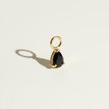 These interchangeable earring charms feature a pear-cut crystal set in a four-prong gold setting, topped with a round jump ring for easy attachment to hoops or huggies. Available in two colorways — a vivid emerald green and a sparkling clear crystal — the charms let shoppers customize and mix up their earring stack with a single pair of base hoops. Photographed flat against a clean, neutral background, the dramatic side lighting highlights each stone's faceted cut and the polished gold prongs, along with th