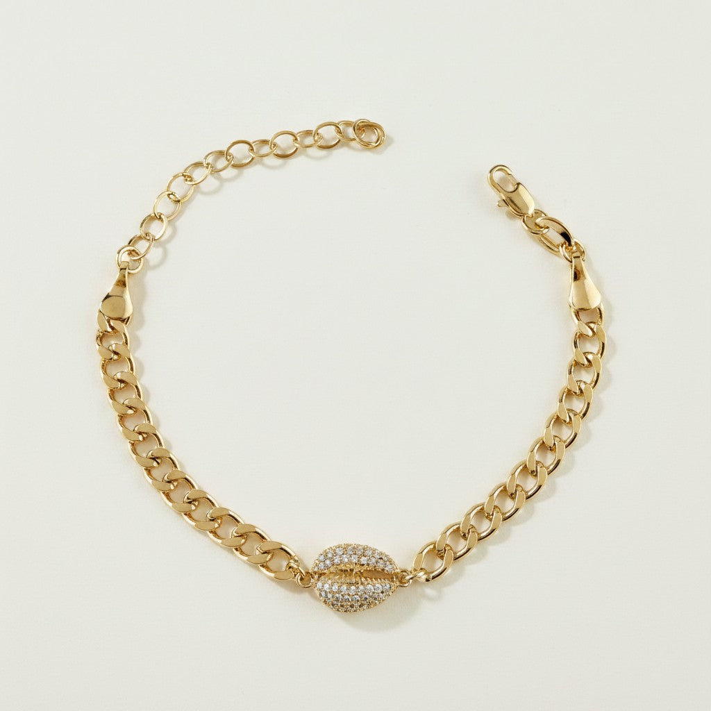 This gold curb chain bracelet pairs a bold, flat-link chain with a pavé crystal cowrie shell charm at the center, creating a coordinated statement piece that matches the cowrie shell choker in the same collection. The chunky curb links offer a substantial, textured base while the crystal-encrusted shell adds sparkle and dimension as the eye-catching focal point. The bracelet fastens with a secure lobster clasp closure and includes a chain extender, allowing for an adjustable, customizable fit around the wri