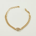 This gold curb chain bracelet pairs a bold, flat-link chain with a pavé crystal cowrie shell charm at the center, creating a coordinated statement piece that matches the cowrie shell choker in the same collection. The chunky curb links offer a substantial, textured base while the crystal-encrusted shell adds sparkle and dimension as the eye-catching focal point. The bracelet fastens with a secure lobster clasp closure and includes a chain extender, allowing for an adjustable, customizable fit around the wri