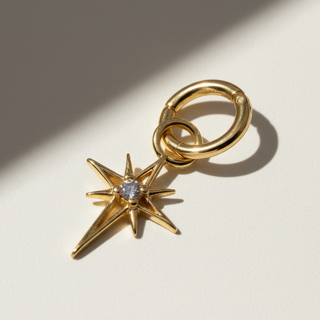 This gold seamless hoop earring is styled with a dazzling eight-point north star charm, featuring a sparkling CZ center stone set in a prong setting at the heart of the star's radiating points. The smooth, hinged hoop clicks open and closed with ease, while the star charm attaches via its own jump ring for a customizable, celestial-inspired look. Photographed at a close angle against a neutral background, dramatic diagonal shadows echo the star's sharp points, highlighting the charm's dimensional, faceted d