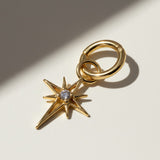 This gold seamless hoop earring is styled with a dazzling eight-point north star charm, featuring a sparkling CZ center stone set in a prong setting at the heart of the star's radiating points. The smooth, hinged hoop clicks open and closed with ease, while the star charm attaches via its own jump ring for a customizable, celestial-inspired look. Photographed at a close angle against a neutral background, dramatic diagonal shadows echo the star's sharp points, highlighting the charm's dimensional, faceted d