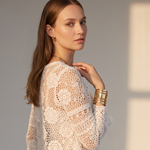 A woman wearing a white crochet lace long-sleeve top and white pants, photographed in warm side lighting against a light gray background.