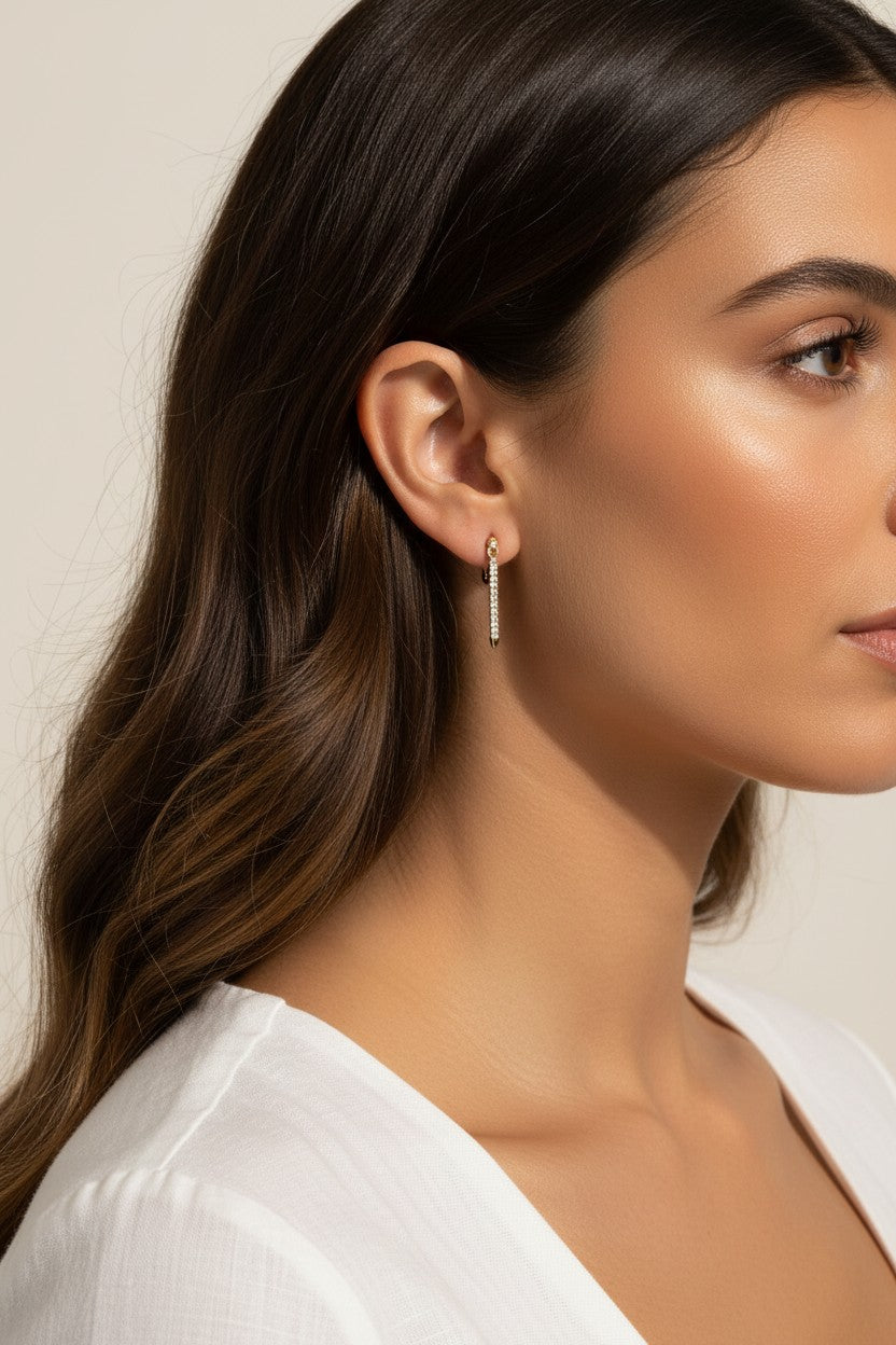 This close-up profile shot shows the longest gold dagger earring worn at the lobe, its tapered pavé crystal spike catching soft light as it extends elegantly downward. Styled with sleek, side-swept waves and a draped white top, the earring is shown at true scale against the jawline, giving shoppers a clear sense of its elongated drop length and sleek, minimal profile. 