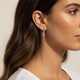 This close-up profile shot shows the longest gold dagger earring worn at the lobe, its tapered pavé crystal spike catching soft light as it extends elegantly downward. Styled with sleek, side-swept waves and a draped white top, the earring is shown at true scale against the jawline, giving shoppers a clear sense of its elongated drop length and sleek, minimal profile. 