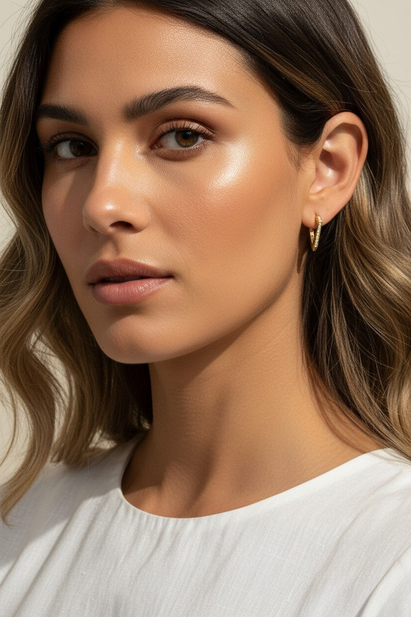 A close-up portrait of a woman wearing a delicate gold huggie hoop earring, shown from the chest up against a soft beige background.