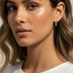 A close-up portrait of a woman wearing a delicate gold huggie hoop earring, shown from the chest up against a soft beige background.
