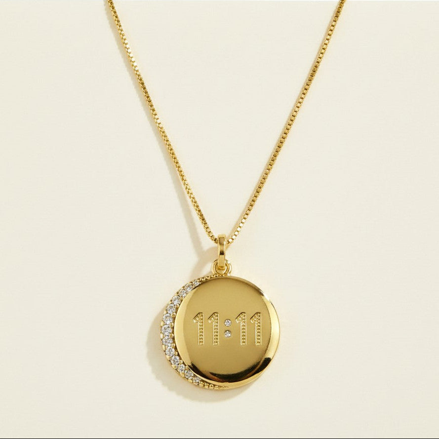 This meaningful gold pendant necklace features a polished round coin charm engraved with the angel number "11:11," a symbol widely associated with intuition, manifestation, and spiritual alignment. A pavé crystal crescent moon overlays one edge of the disc, adding sparkle and celestial symbolism while framing the engraved numerals with a single bezel-set crystal accent between the digits. The pendant hangs from a delicate box chain and bail, creating a clean, layerable silhouette perfect for everyday wear o