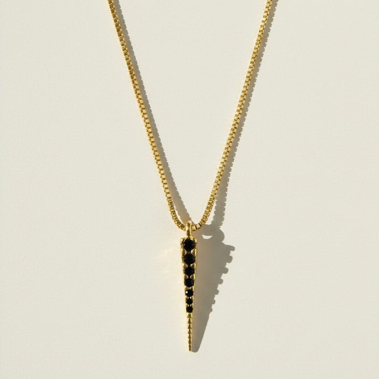 This gold necklace features a sleek dagger-shaped pendant lined with sparkling black pavé crystals, tapering from a wider top edge down to a smooth, polished point for a bold, edgy silhouette. The pendant hangs from a fine box chain, creating a clean, versatile drape that layers effortlessly with other necklaces