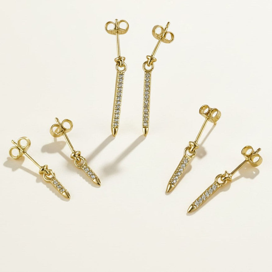 This set showcases a gold dagger-shaped drop earring in three graduated lengths, each featuring a tapered spike lined with sparkling pavé crystals that narrows to a smooth, polished point. The elongated silhouette hangs from a secure post-and-butterfly back closure, offering a sleek, edgy alternative to traditional studs. Photographed flat against a clean, neutral background, the trio displays the earrings' varying drop lengths side by side.