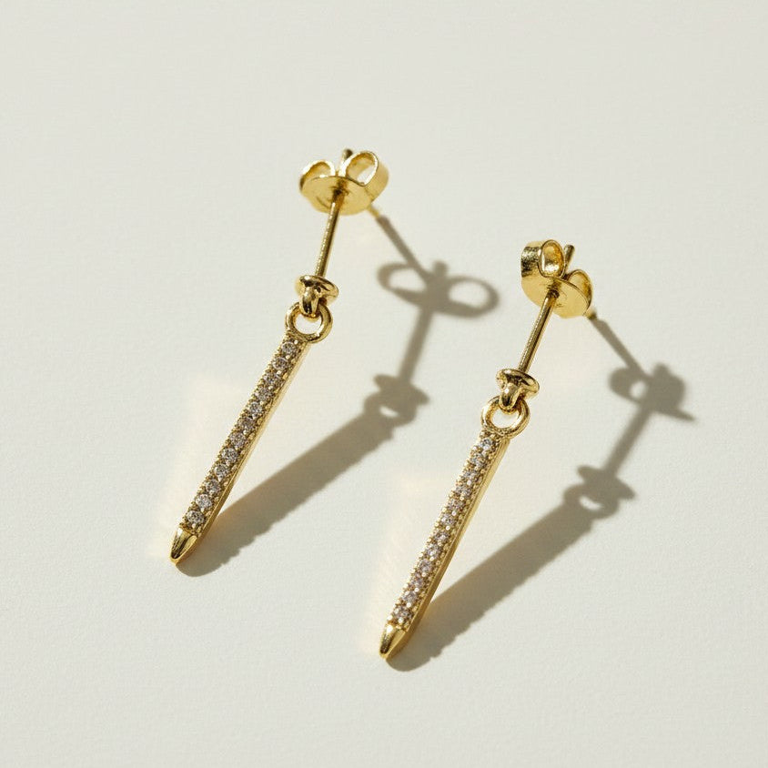 This set showcases a gold dagger-shaped drop earring in three graduated lengths, each featuring a tapered spike lined with sparkling pavé crystals that narrows to a smooth, polished point. The elongated silhouette hangs from a secure post-and-butterfly back closure, offering a sleek, edgy alternative to traditional studs. Photographed flat against a clean, neutral background, the trio displays the earrings' varying drop lengths side by side, highlighting the fine crystal detailing.