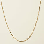This delicate gold necklace is crafted from a continuous row of tiny flat sequin-like discs linked edge to edge, creating a shimmering, textured chain that catches the light with every angle. The fine, low-profile links lie smooth against the skin, offering a subtle sparkle that pairs beautifully for layering or wearing alone. It fastens securely with a lobster clasp closure and includes a short oval-link extender chain with a polished ball-chain detail, allowing for an adjustable, customizable fit.