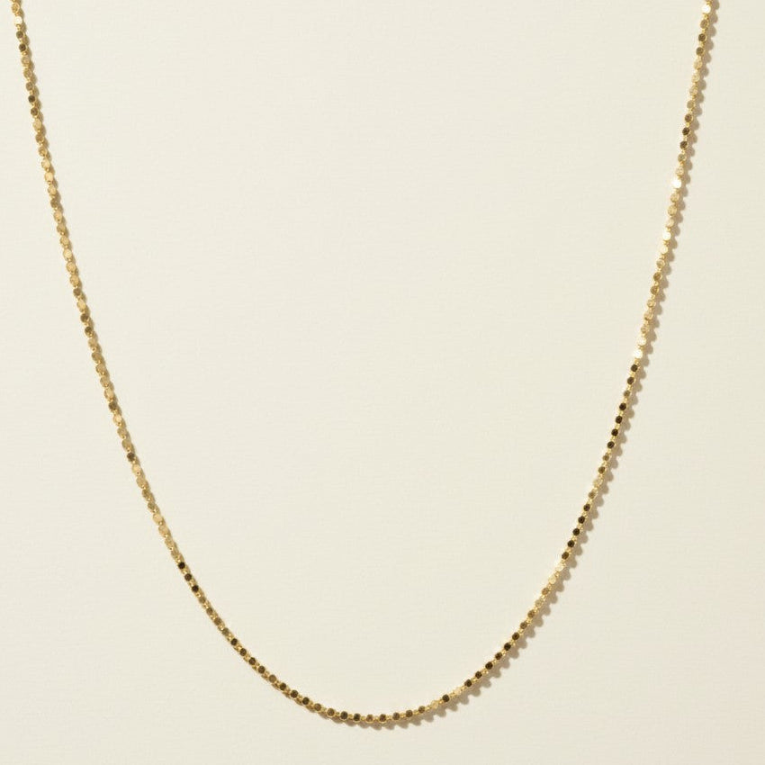 This delicate gold necklace is crafted from a continuous row of tiny flat sequin-like discs linked edge to edge, creating a shimmering, textured chain that catches the light with every angle. The fine, low-profile links lie smooth against the skin, offering a subtle sparkle that pairs beautifully for layering or wearing alone. It fastens securely with a lobster clasp closure and includes a short oval-link extender chain with a polished ball-chain detail, allowing for an adjustable, customizable fit.