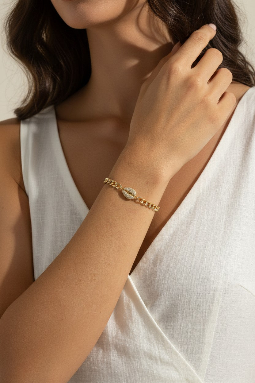 This close-up lifestyle shot shows a gold curb chain bracelet resting elegantly on the wrist, with the pavé crystal cowrie shell charm sitting front and center as the piece's focal point. Styled with a white linen wrap top, the bracelet's bold chain links and sparkling shell charm are shown at true scale against warm skin tones, giving shoppers a clear sense of fit and drape. Soft, warm natural lighting and a relaxed hand-to-collarbone pose lend the image an intimate, editorial feel, highlighting the interp