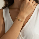 This close-up lifestyle shot shows a gold curb chain bracelet resting elegantly on the wrist, with the pavé crystal cowrie shell charm sitting front and center as the piece's focal point. Styled with a white linen wrap top, the bracelet's bold chain links and sparkling shell charm are shown at true scale against warm skin tones, giving shoppers a clear sense of fit and drape. Soft, warm natural lighting and a relaxed hand-to-collarbone pose lend the image an intimate, editorial feel, highlighting the interp