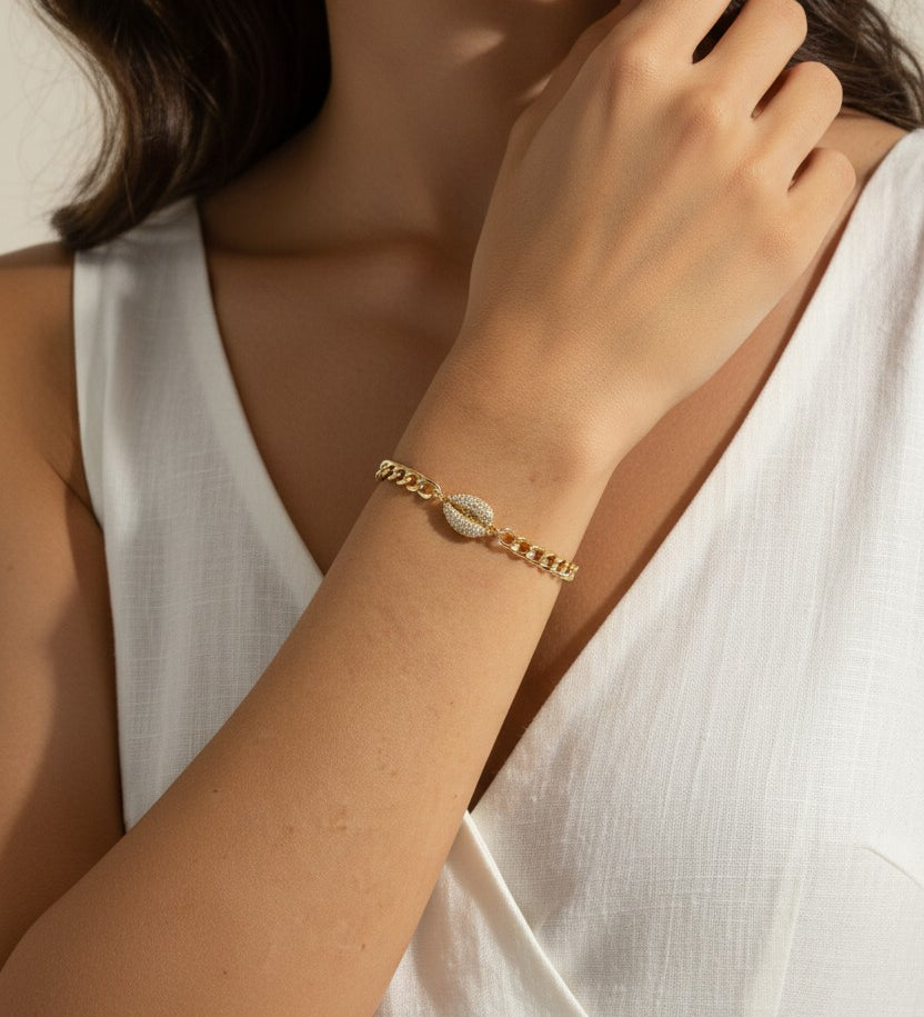 This close-up lifestyle shot shows a gold curb chain bracelet resting elegantly on the wrist, with the pavé crystal cowrie shell charm sitting front and center as the piece's focal point. Styled with a white linen wrap top, the bracelet's bold chain links and sparkling shell charm are shown at true scale against warm skin tones, giving shoppers a clear sense of fit and drape. Soft, warm natural lighting and a relaxed hand-to-collarbone pose lend the image an intimate, editorial feel, highlighting the interp