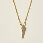 This gold necklace features a sleek dagger-shaped pendant lined with sparkling pavé crystals, tapering from a wider top edge down to a smooth, polished point for a sharp, minimalist silhouette. The pendant hangs from a fine box chain, creating a clean, versatile drape that layers effortlessly with other necklaces.