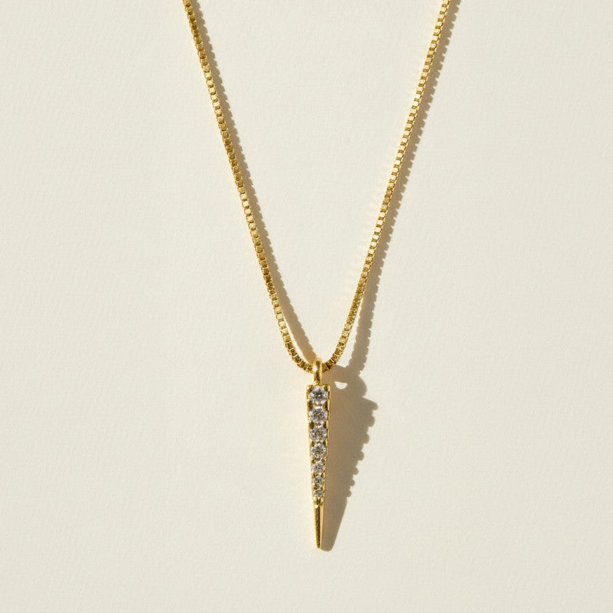 This gold necklace features a sleek dagger-shaped pendant lined with sparkling pavé crystals, tapering from a wider top edge down to a smooth, polished point for a sharp, minimalist silhouette. The pendant hangs from a fine box chain, creating a clean, versatile drape that layers effortlessly with other necklaces.