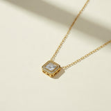 This gold-tone paperclip chain necklace is centered on a square-cut pendant with a large clear crystal set in a polished bezel. A pavé halo of tiny clear stones surrounds the center stone and adds sparkle at every angle. The first frame is a straight-on flat lay on a warm cream surface, showing the full pendant face and the pavé border. The second frame is an angled close-up of the pendant’s slotted, gridded side walls and the pavé setting. The third frame is a detail of the clasp, a lobster clasp joined to