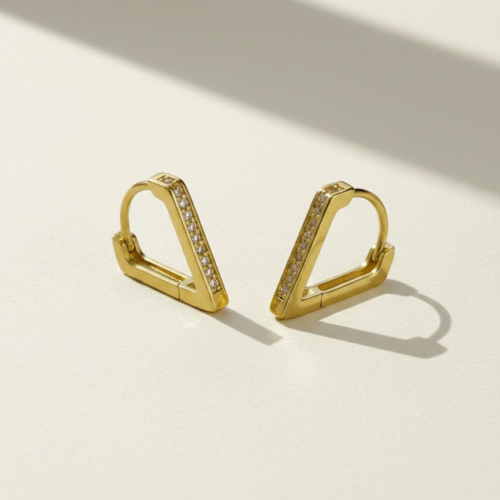 A pair of gold geometric hoop earrings with a row of small clear stones, displayed on a light beige surface with soft shadows.