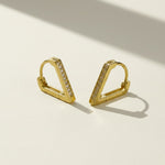 A pair of gold geometric hoop earrings with a row of small clear stones, displayed on a light beige surface with soft shadows.