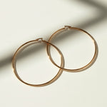 This pair of large gold hoop earrings features a smooth, uninterrupted circular silhouette crafted from a sleek round wire, offering a timeless, versatile shape that works for both everyday wear and dressed-up occasions. The hoops close securely with a hinged snap-back mechanism, ensuring a comfortable, secure fit at the earlobe. 