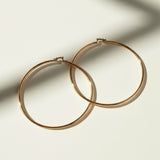 This pair of large gold hoop earrings features a smooth, uninterrupted circular silhouette crafted from a sleek round wire, offering a timeless, versatile shape that works for both everyday wear and dressed-up occasions. The hoops close securely with a hinged snap-back mechanism, ensuring a comfortable, secure fit at the earlobe. 