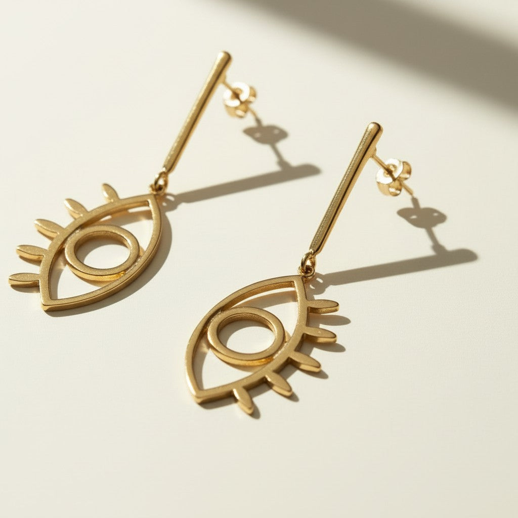 evil eye drop earrings, gold cutout earrings, matte gold dangle earrings, symbolic jewelry, geometric drop earrings, minimalist gold earrings, statement drop earrings flat lay, open linework earrings