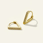 This pair of gold huggie earrings features a bold, architectural triangle silhouette with sharp, angular edges for a modern, geometric look. One flat edge of each triangle is lined with a row of sparkling pavé-set crystals, adding a subtle sparkle that catches the light while the smooth gold planes provide clean, minimalist contrast. Designed as a huggie hoop, the earrings hinge closed with a click-top closure for a secure, comfortable fit that hugs the earlobe. Photographed at an angle against a warm neutr