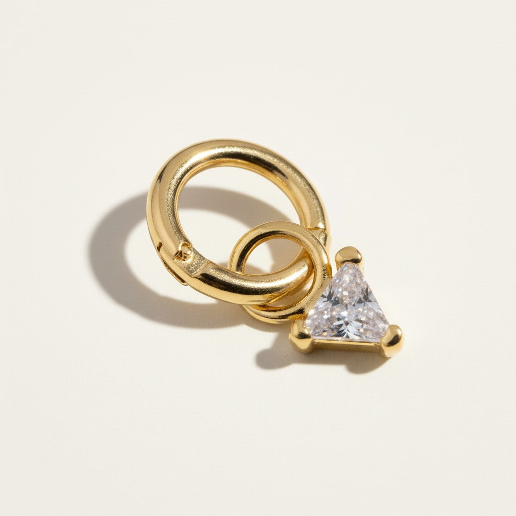 This gold seamless hoop earring is styled with a sparkling trillion-cut CZ charm, its bold triangular stone set in a sleek three-prong gold setting for a modern, geometric accent. The smooth, hinged hoop clicks open and closed with ease, while the charm attaches via its own jump ring for a customizable, interchangeable look. Photographed at a close angle against a neutral background, warm directional lighting catches the facets of the trillion-cut stone, highlighting its clean angular lines and brilliant sp