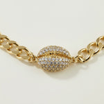 This gold curb chain bracelet pairs a bold, flat-link chain with a pavé crystal cowrie shell charm at the center, creating a coordinated statement piece that matches the cowrie shell choker in the same collection. The chunky curb links offer a substantial, textured base while the crystal-encrusted shell adds sparkle and dimension as the eye-catching focal point