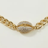 This gold curb chain bracelet pairs a bold, flat-link chain with a pavé crystal cowrie shell charm at the center, creating a coordinated statement piece that matches the cowrie shell choker in the same collection. The chunky curb links offer a substantial, textured base while the crystal-encrusted shell adds sparkle and dimension as the eye-catching focal point