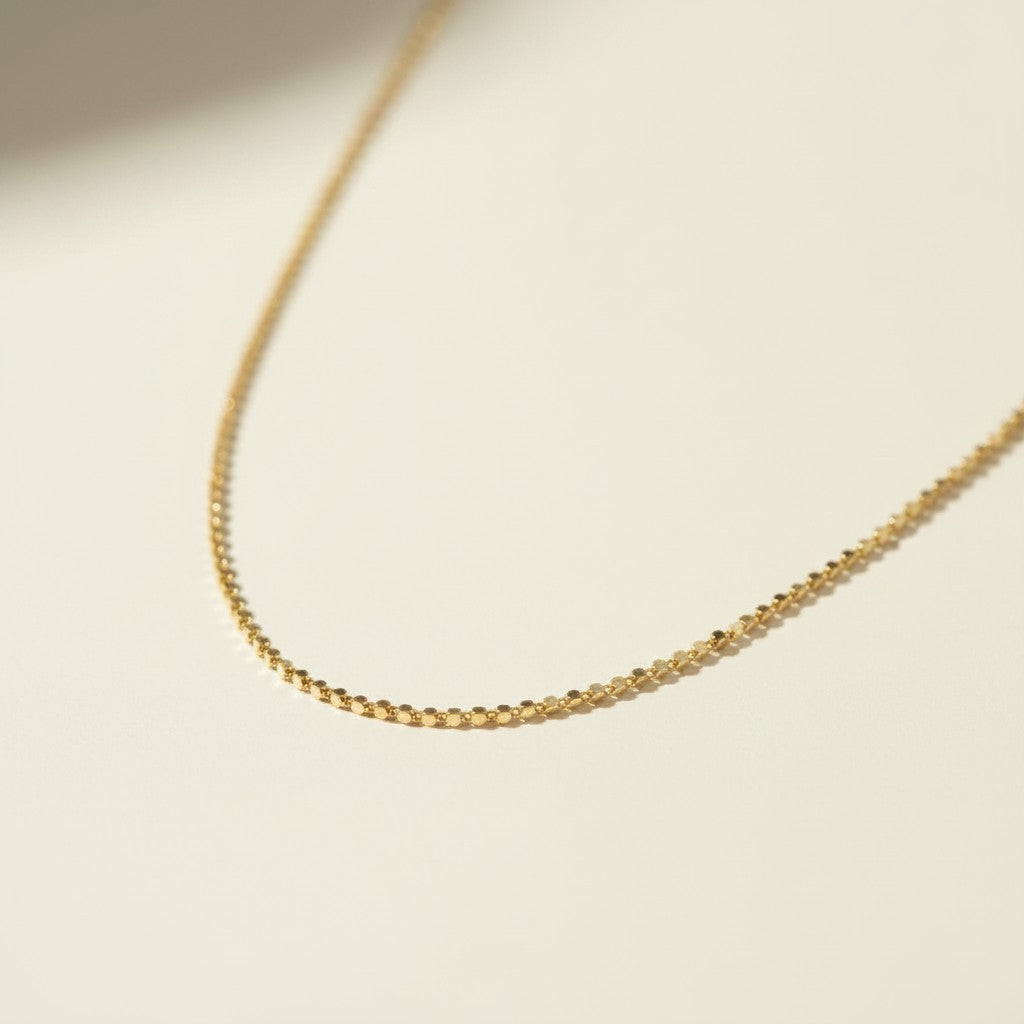 This delicate gold necklace is crafted from a continuous row of tiny flat sequin-like discs linked edge to edge, creating a shimmering, textured chain that catches the light with every angle. The fine, low-profile links lie smooth against the skin, offering a subtle sparkle that pairs beautifully for layering or wearing alone. It fastens securely with a lobster clasp closure and includes a short oval-link extender chain with a polished ball-chain detail, allowing for an adjustable, customizable fit.