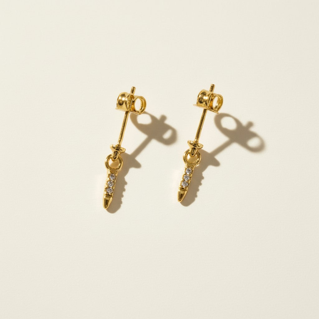 This set showcases a gold dagger-shaped drop earring in three graduated lengths, each featuring a tapered spike lined with sparkling pavé crystals that narrows to a smooth, polished point. The elongated silhouette hangs from a secure post-and-butterfly back closure, offering a sleek, edgy alternative to traditional studs. Photographed flat against a clean, neutral background, the trio displays the earrings' varying drop lengths side by side, highlighting the fine crystal detailing.