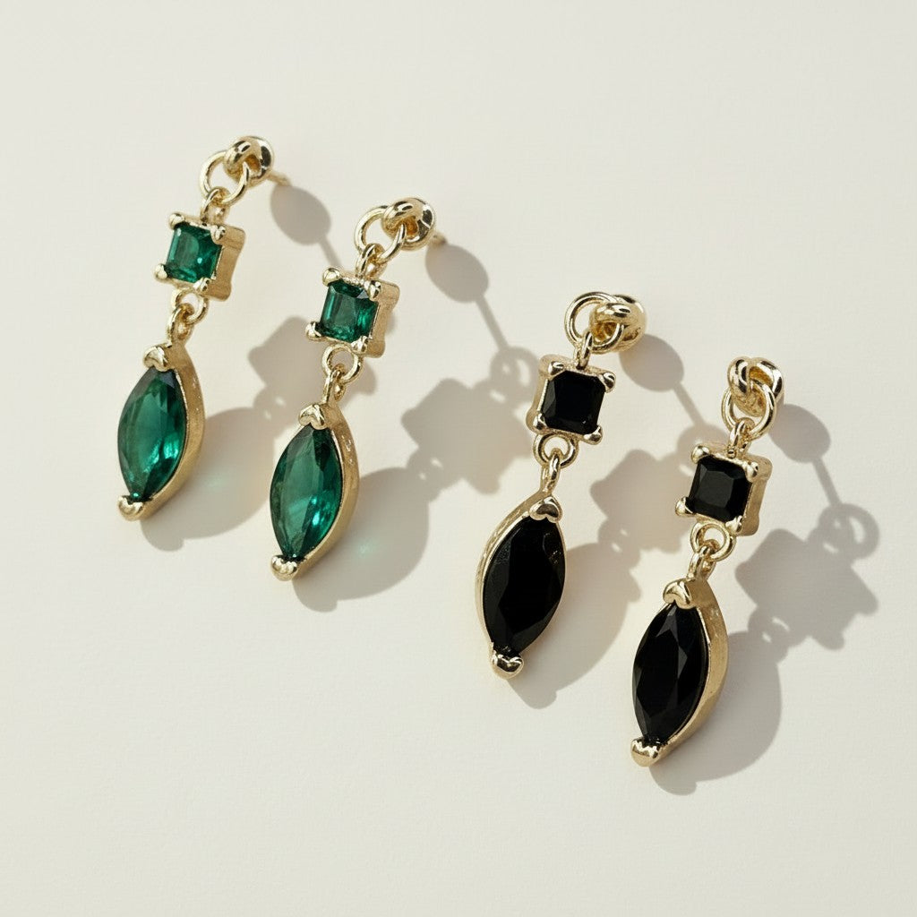 This pair of gold dangle earrings features a two-tier design combining a square-cut crystal stud with a faceted marquise-cut stone drop below, set in polished gold prong settings. Shown in two striking colorways — a rich emerald green and a sleek jet black — the earrings offer a versatile statement piece that transitions easily from daytime to evening wear. Photographed flat against a clean, neutral background with warm directional lighting, the dramatic shadow play highlights the faceted cut and dimensiona