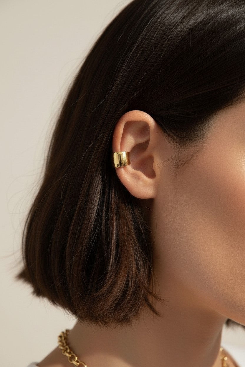 This close-up profile shot shows a chunky gold ear cuff wrapped securely around the upper ear, its smooth domed silhouette catching soft light for a warm, polished shine. Styled against sleek dark hair and paired with a glimpse of a layered gold chain necklace, the cuff is shown at true scale, giving shoppers a clear sense of its bold, wide-band fit and no-piercing-required design. The tight crop and soft, warm lighting create a clean, editorial beauty shot, highlighting how the sculptural gold piece sits c