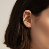 This close-up profile shot shows a chunky gold ear cuff wrapped securely around the upper ear, its smooth domed silhouette catching soft light for a warm, polished shine. Styled against sleek dark hair and paired with a glimpse of a layered gold chain necklace, the cuff is shown at true scale, giving shoppers a clear sense of its bold, wide-band fit and no-piercing-required design. The tight crop and soft, warm lighting create a clean, editorial beauty shot, highlighting how the sculptural gold piece sits c