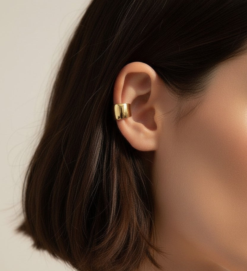 This close-up profile shot shows a chunky gold ear cuff wrapped securely around the upper ear, its smooth domed silhouette catching soft light for a warm, polished shine. Styled against sleek dark hair and paired with a glimpse of a layered gold chain necklace, the cuff is shown at true scale, giving shoppers a clear sense of its bold, wide-band fit and no-piercing-required design. The tight crop and soft, warm lighting create a clean, editorial beauty shot, highlighting how the sculptural gold piece sits c