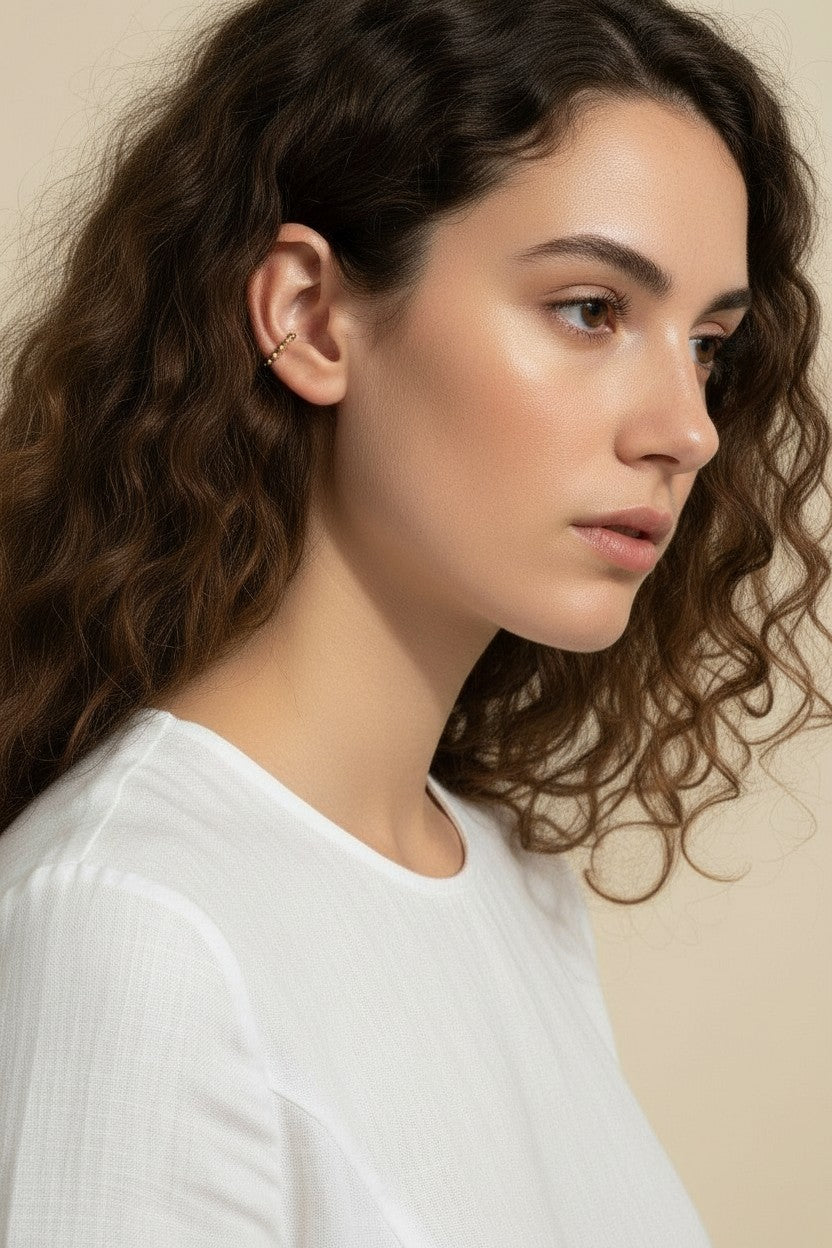 This Gold Beaded Ear Cuff takes a minimalist approach to ear styling with a delicate row of small polished gold beads curving along the upper cartilage. Shown here in close-up profile on a model with soft curls and a relaxed white top, the cuff sits neatly at the top of the ear for a subtle, no-piercing accent that layers beautifully with studs or hoops. Its slim