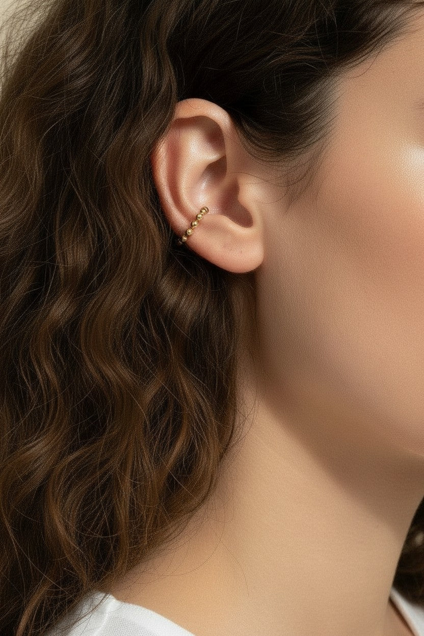 This Gold Beaded Ear Cuff takes a minimalist approach to ear styling with a delicate row of small polished gold beads curving along the upper cartilage. Shown here in close-up profile on a model with soft curls and a relaxed white top, the cuff sits neatly at the top of the ear for a subtle, no-piercing accent that layers beautifully with studs or hoops. Its slim