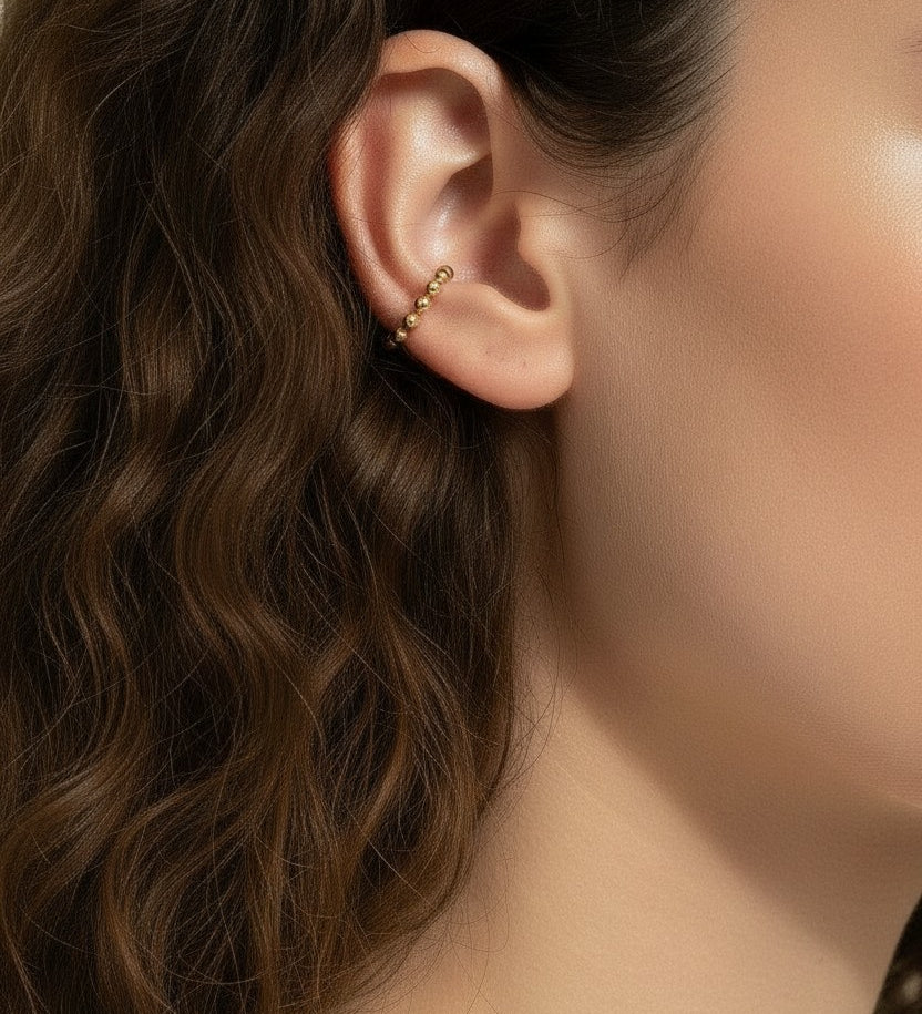 This Gold Beaded Ear Cuff takes a minimalist approach to ear styling with a delicate row of small polished gold beads curving along the upper cartilage. Shown here in close-up profile on a model with soft curls and a relaxed white top, the cuff sits neatly at the top of the ear for a subtle, no-piercing accent that layers beautifully with studs or hoops. Its slim
