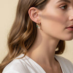 This close-up profile shot shows a pair of small gold hoop earrings, each accented with a dangling pear-cut crystal charm — one in emerald green, one in clear crystal — worn together along the lower earlobe. The delicate hoops sit close to the ear with the faceted charms swinging just below, shown at true scale against soft waved hair and a white wrap top for a clear sense of drop length and fit. Warm, soft lighting creates a clean, romantic mood, highlighting how the mixed emerald-and-clear pairing offers 