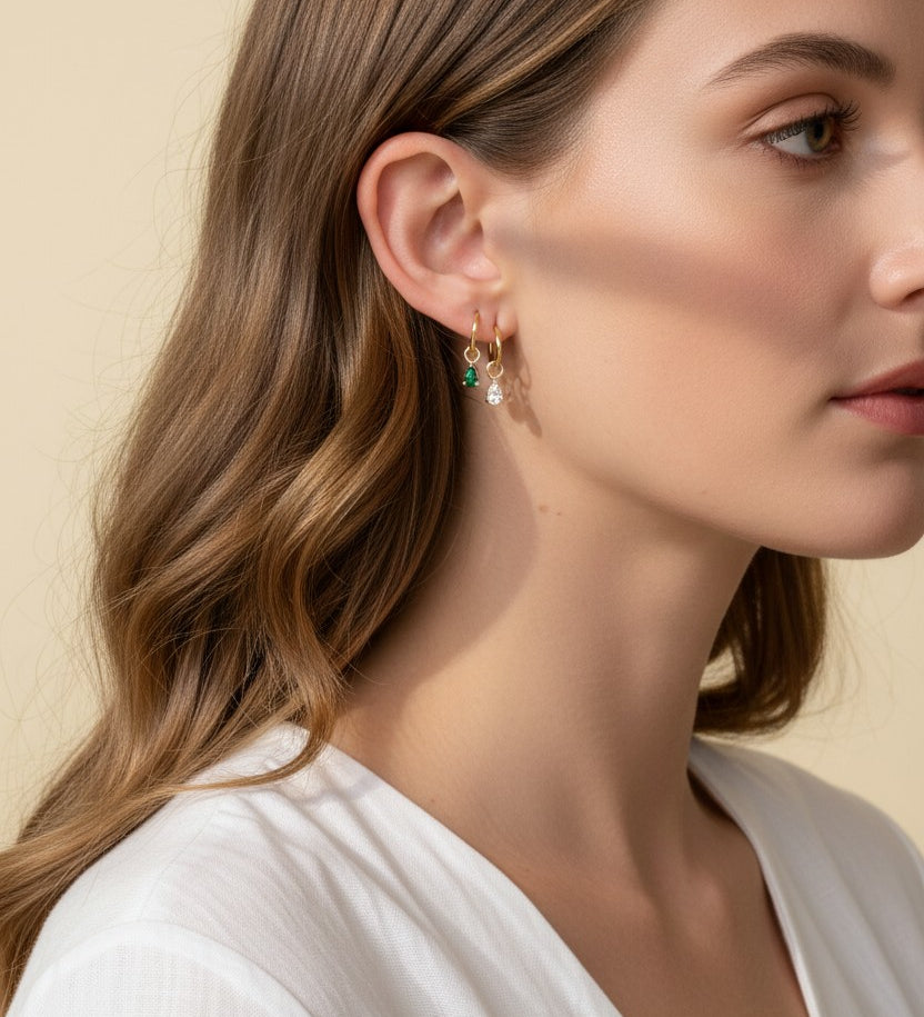 This close-up profile shot shows a pair of small gold hoop earrings, each accented with a dangling pear-cut crystal charm — one in emerald green, one in clear crystal — worn together along the lower earlobe. The delicate hoops sit close to the ear with the faceted charms swinging just below, shown at true scale against soft waved hair and a white wrap top for a clear sense of drop length and fit. Warm, soft lighting creates a clean, romantic mood, highlighting how the mixed emerald-and-clear pairing offers 