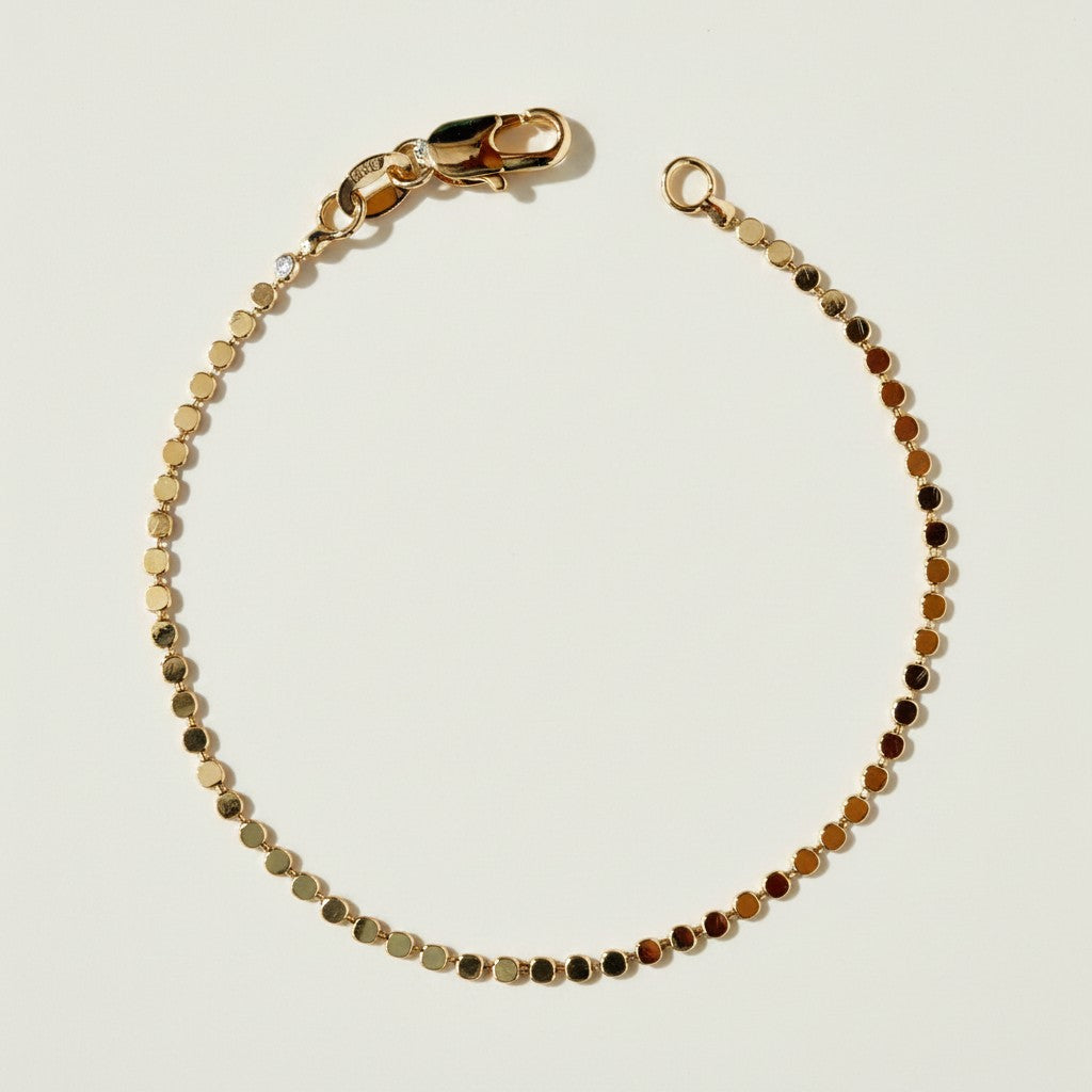 This delicate gold bracelet is crafted from a continuous row of small, flat circular discs linked edge to edge, creating a smooth, coin-like texture that catches the light with every angle. A single sparkling diamond accent is set discreetly near the clasp, adding a subtle touch of shine to the polished gold links. The bracelet fastens securely with a lobster clasp closure, finished with a maker's stamp detail on the clasp tab for an authentic, quality touch. Photographed flat against a clean, neutral backg