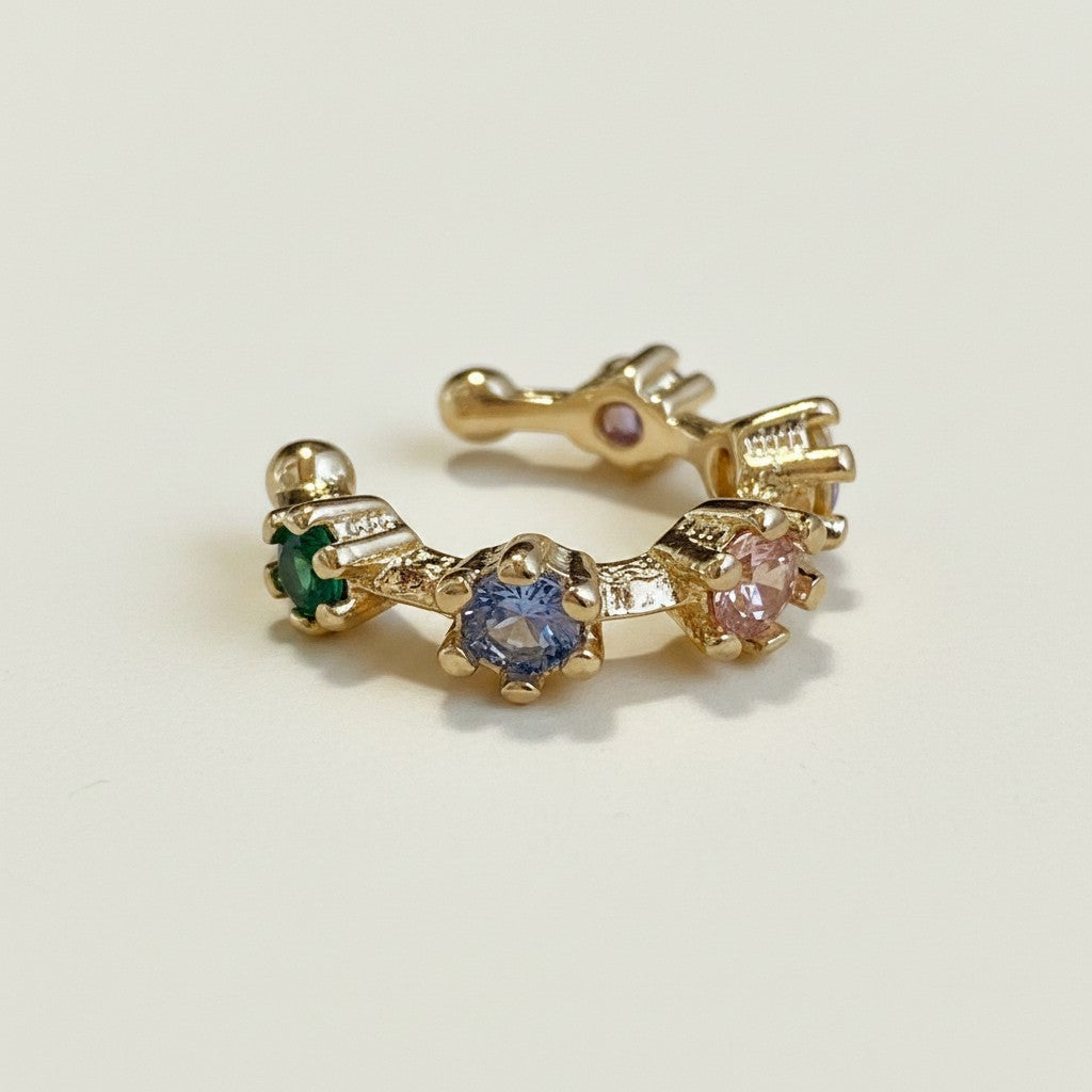 This gold ear cuff features a delicate open band set with a row of multicolor crystals — including emerald green, sapphire blue, and soft pink stones — each held in place with a scalloped prong setting for a dimensional, jeweled effect. The open-back design allows for a flexible, adjustable fit that wraps comfortably around the ear cartilage with no piercing required. Photographed at a close angle against a clean, neutral background, the cuff displays its curved silhouette and the vibrant, rainbow-hued gems